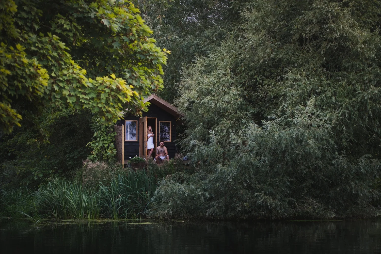 The Secret Sauna, a small black hut half hidden in green riverbank trees